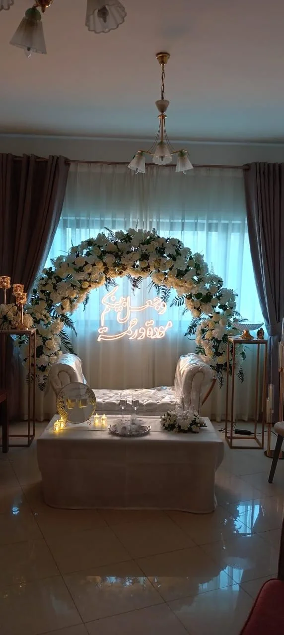 Indoor engagement stage with white floral arch, couple seating and illuminated Arabic script