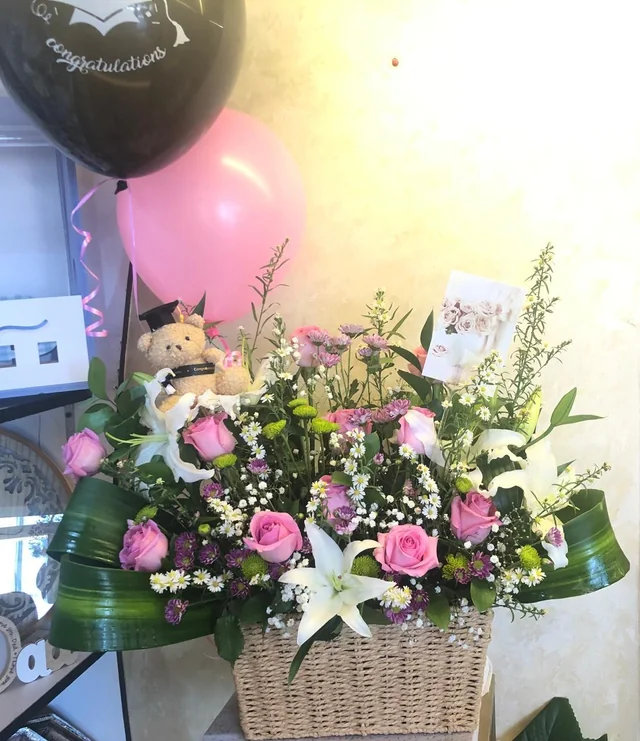Graduation flower basket with pink and white blooms, a graduate teddy and congratulatory balloons