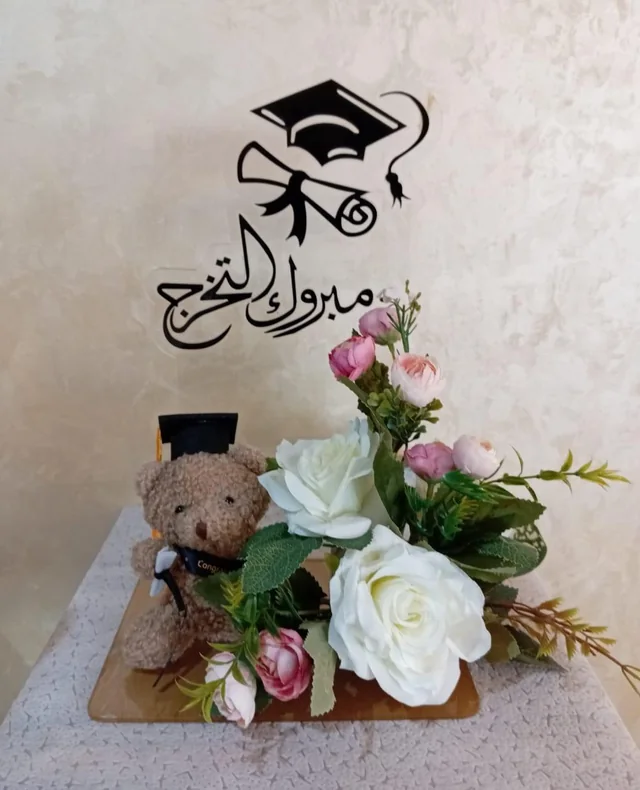 Graduation stand with cap and diploma graphics, Arabic congratulations, flowers and a graduate teddy