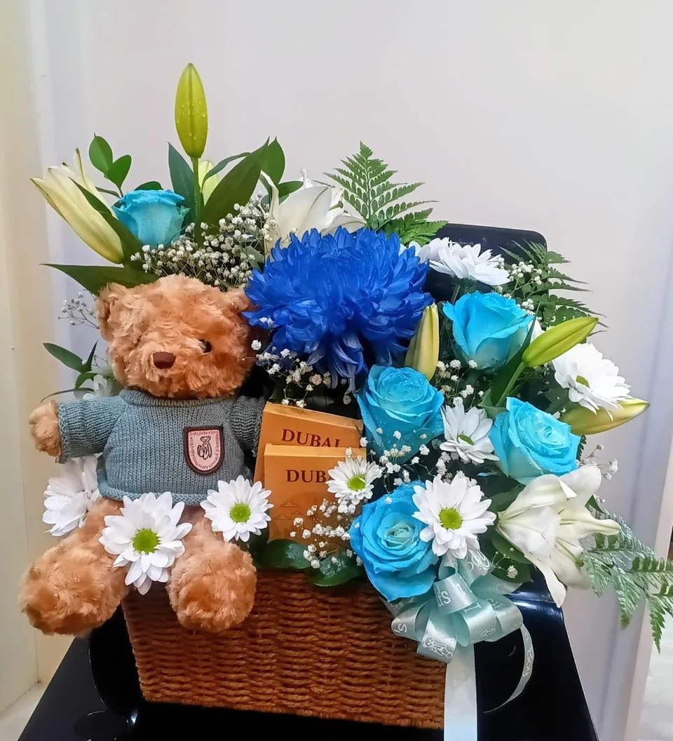 Newborn boy gift basket with blue and white flowers, a teddy bear and chocolates