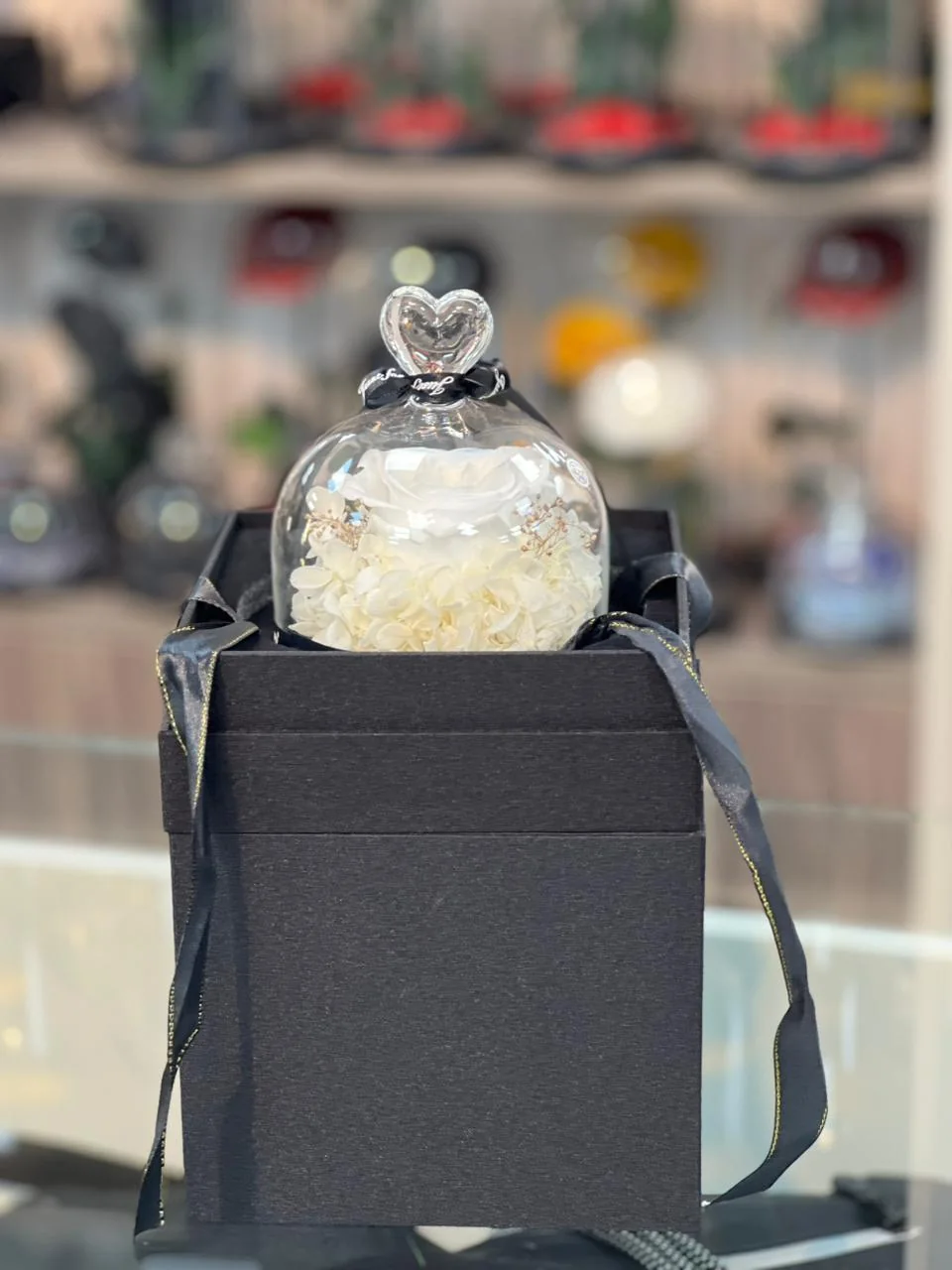 White infinite rose in a small heart-topped glass ornament on a black gift box